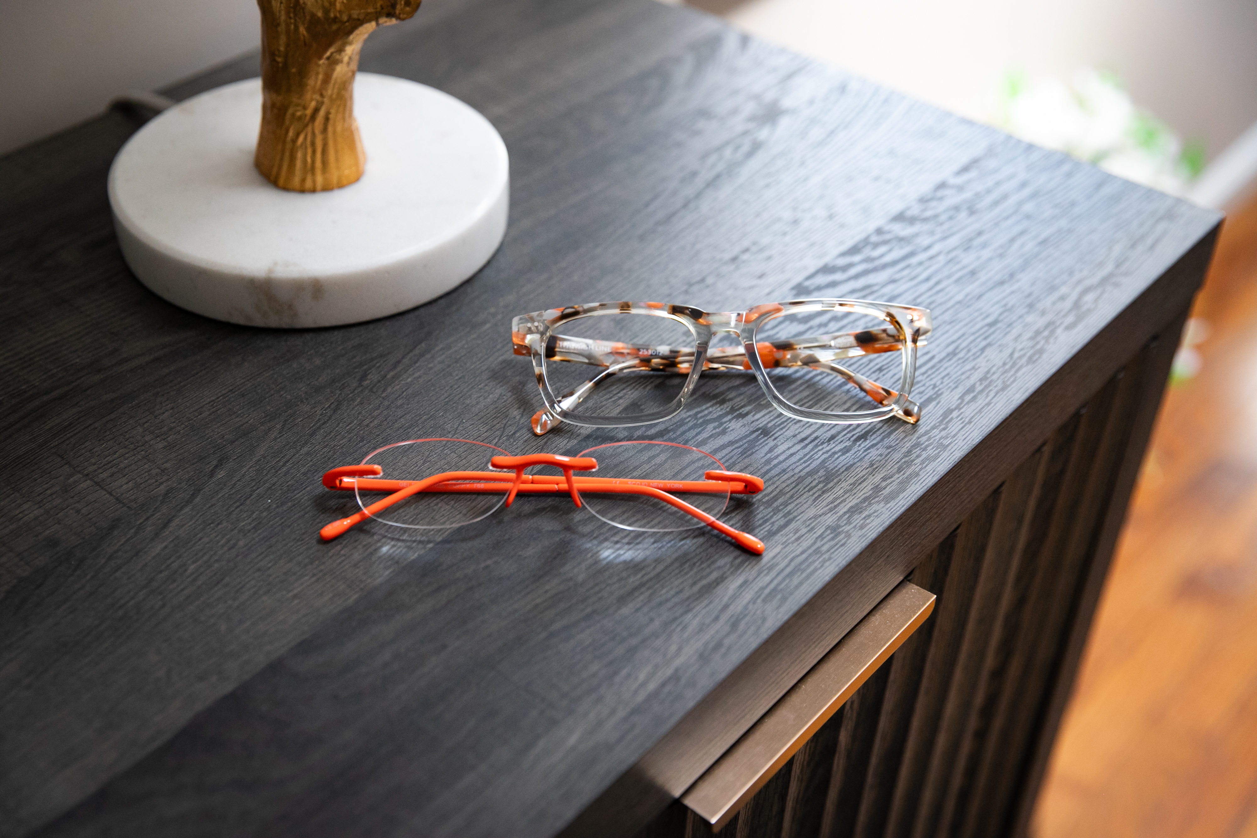Scojo Reading Glasses Two pairs of SCOJO reading glasses ordered online: orange rimless Gels readers and clear tortoise acetate frames on a home sideboard